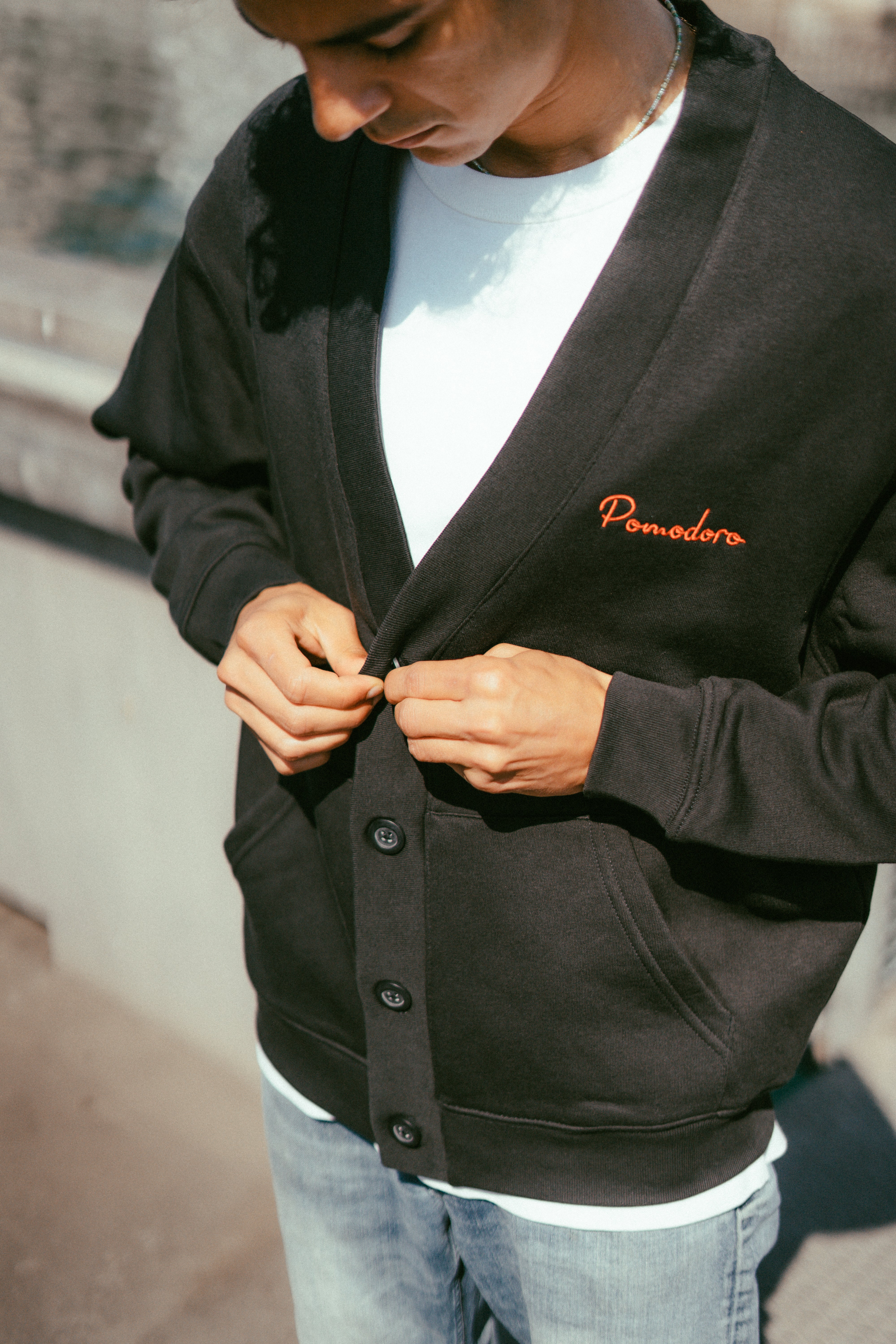 Cardigan noir avec pochettes, unisexe porté par un modèle, coupe oversize avec broderie en rouge, avec logo Pomodoro, côté cœur.