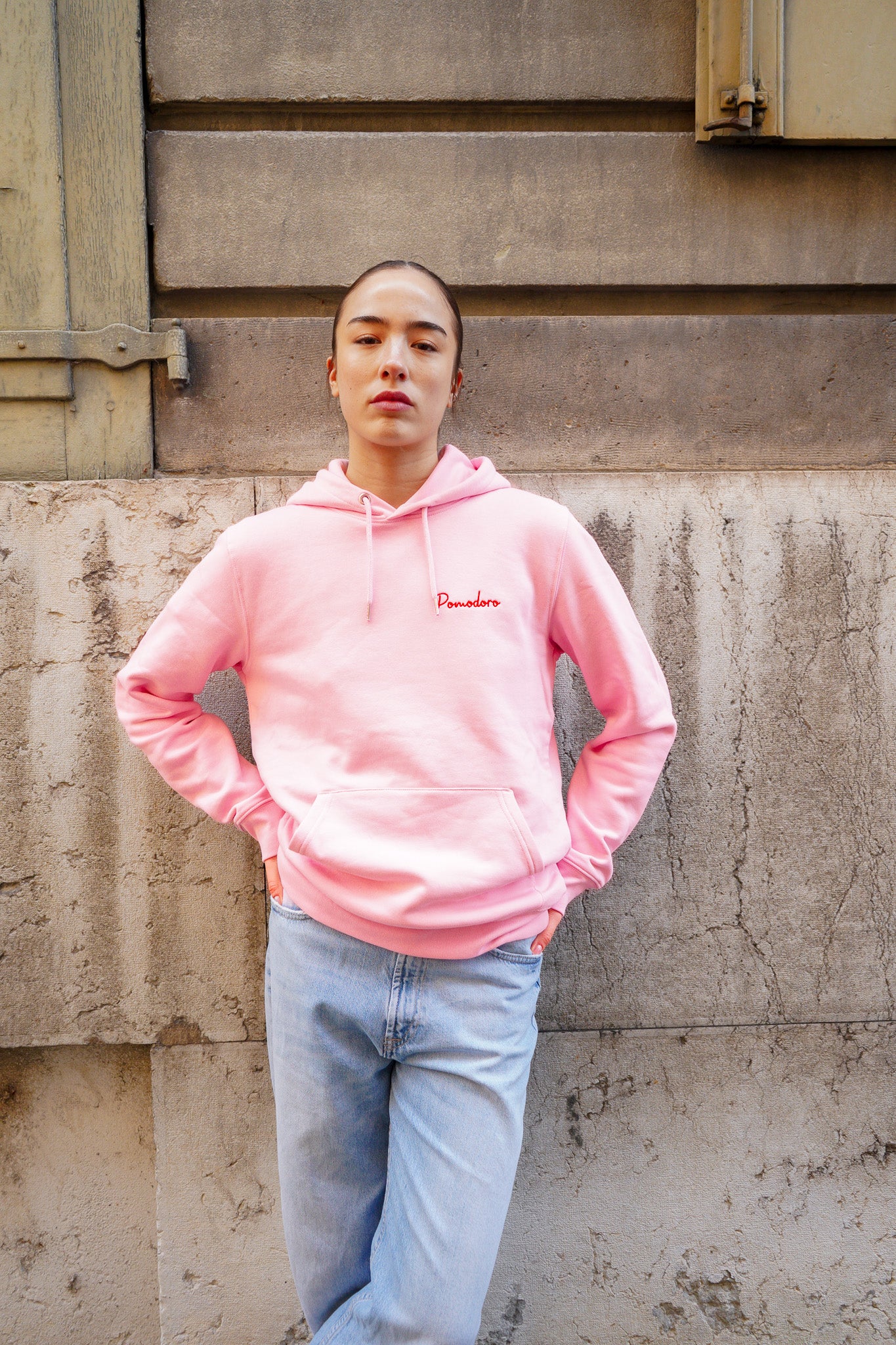 Hoodie rose Rose Rosette Pomodoro en coton bio porté par un modèle debout contre un mur.